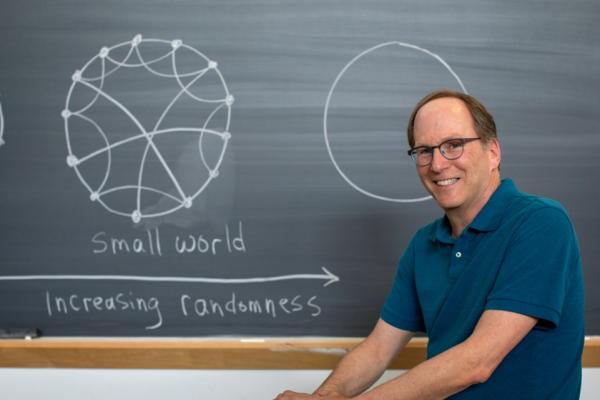 Steven Strogatz in front of a blackboard with "small world" and an illustration on it showing a circle and interconnected lines inside